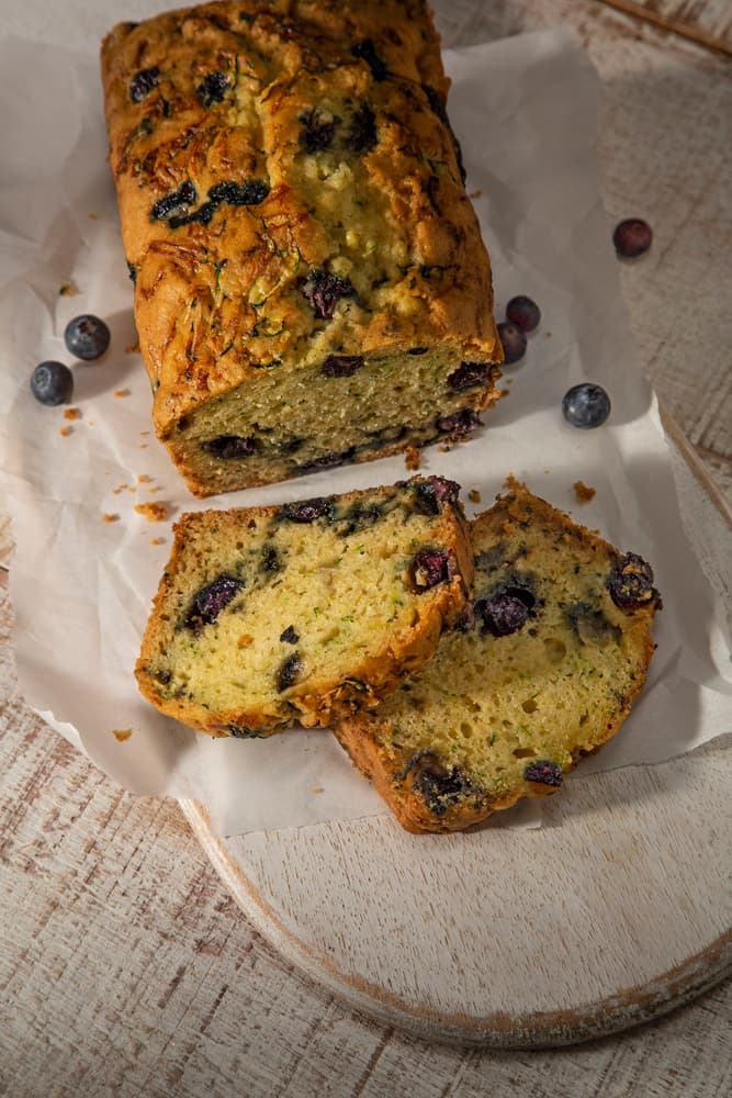 A loaf of blueberry bread with two slices cut, resting on parchment paper atop a wooden board, with a few loose blueberries nearby.