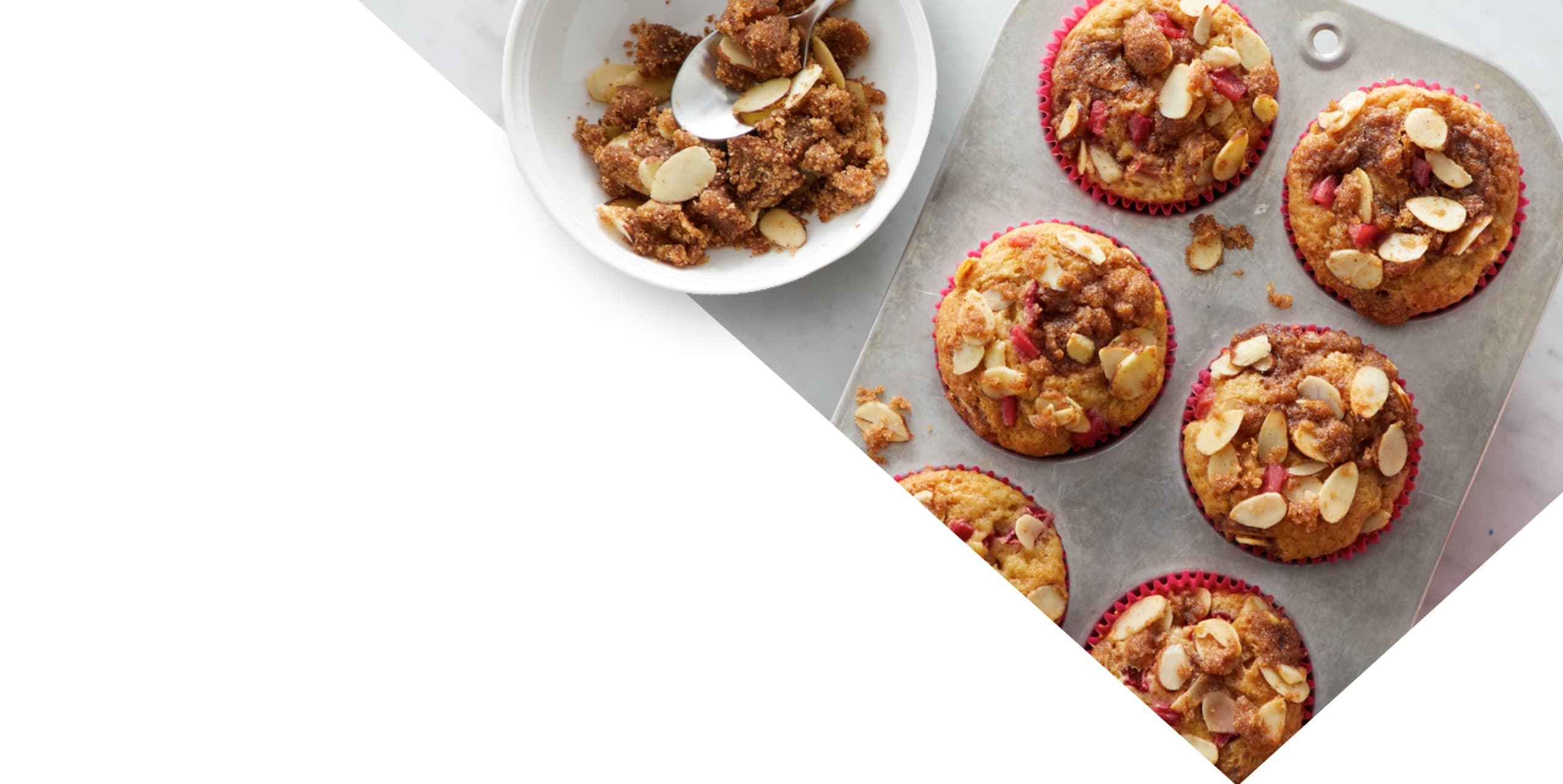 A muffin tray with six baked muffins topped with sliced almonds next to a bowl of streusel mixture and a spoon, on a white surface.