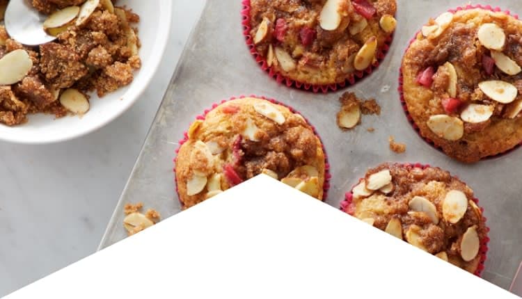 Four muffins topped with sliced almonds and crumbly brown sugar on a baking tray, with a bowl of streusel mixture and a spoon on the side.