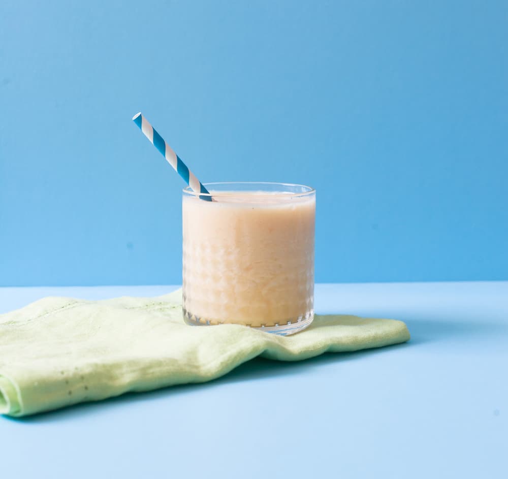 A glass of light orange smoothie with a striped straw sits on a green cloth against a blue background.