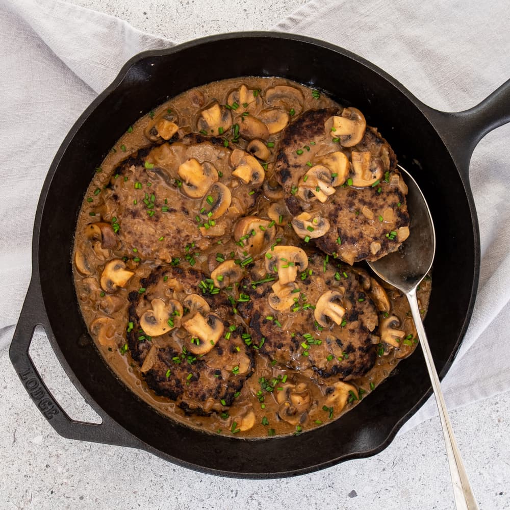 Four hamburger steaks in mushroom gravy are cooked in a cast iron skillet, garnished with chopped chives, with a spoon resting in the pan.