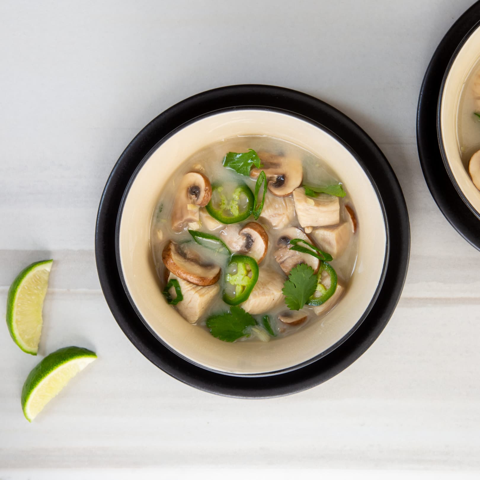 Bowl of soup with chicken, mushrooms, cilantro, and sliced green chili, placed on a white surface next to lime wedges.