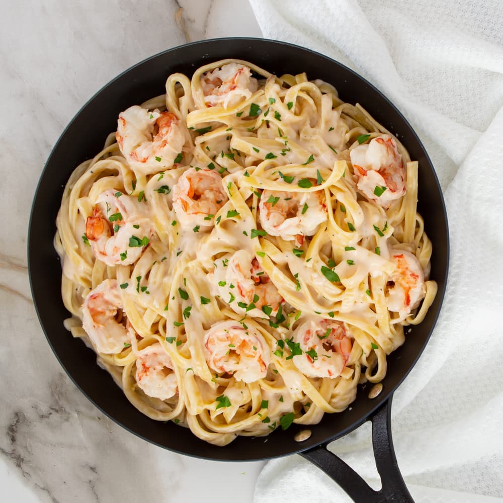 A black skillet filled with fettuccine pasta, shrimp, and creamy white sauce, garnished with chopped parsley, placed on a marble surface with a white towel nearby.