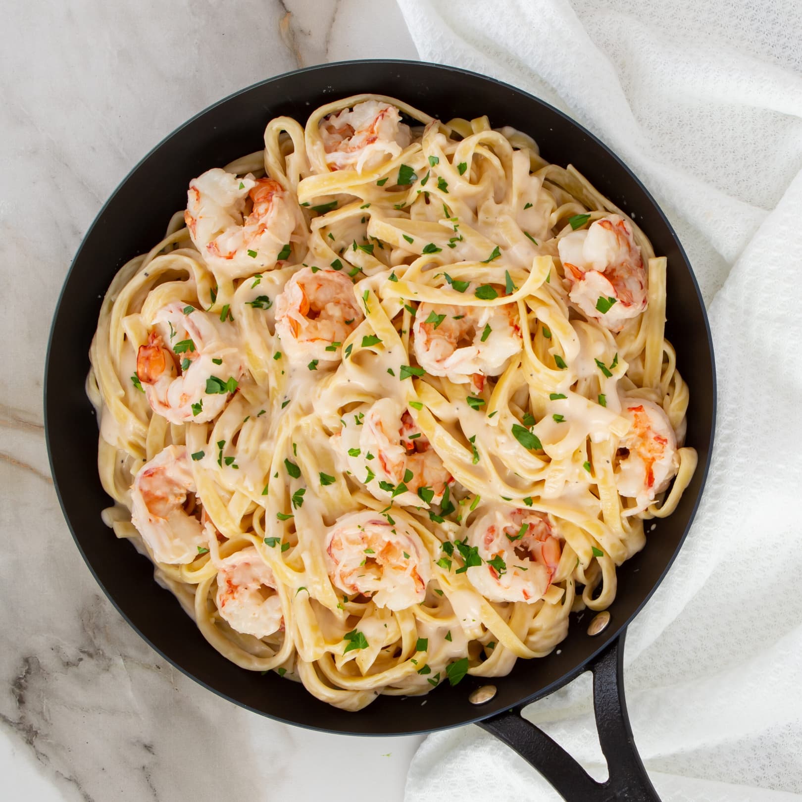 A black skillet filled with fettuccine pasta, shrimp, and creamy white sauce, garnished with chopped parsley, placed on a marble surface with a white towel nearby.