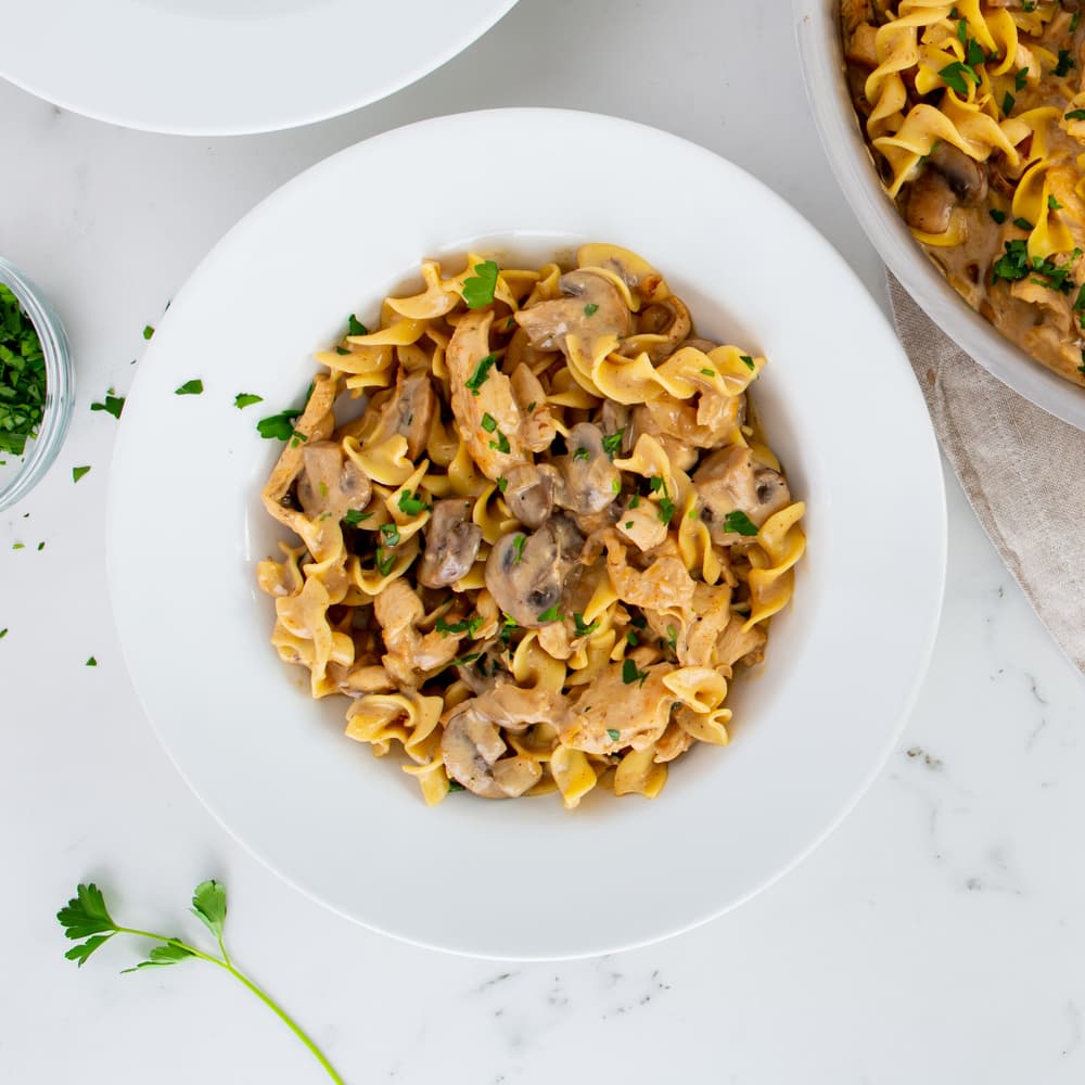 A white bowl filled with egg noodles, creamy sauce, sliced mushrooms, and chopped parsley, sitting on a white marble surface.