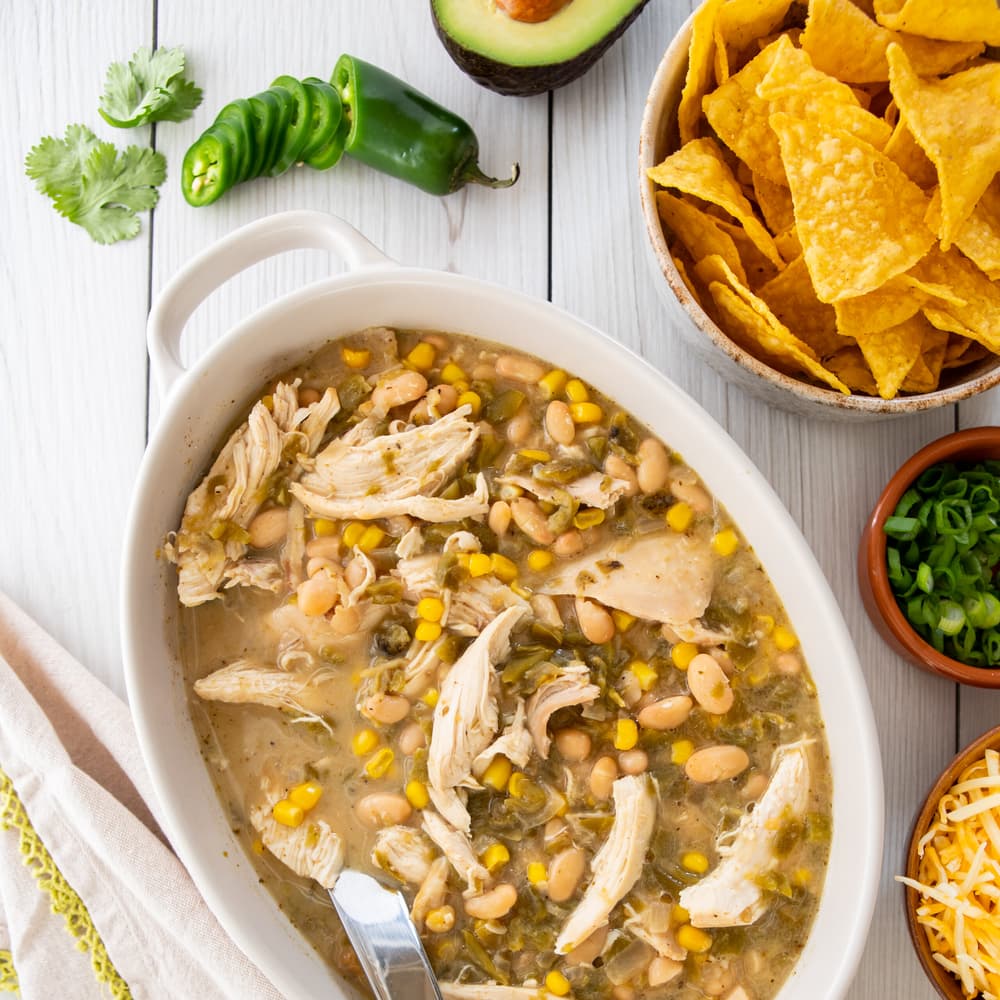 Oval dish of chicken chili with white beans and corn. Surrounding items include tortilla chips, sliced green onion, jalapeño, avocado half, and cilantro on a white wooden table.