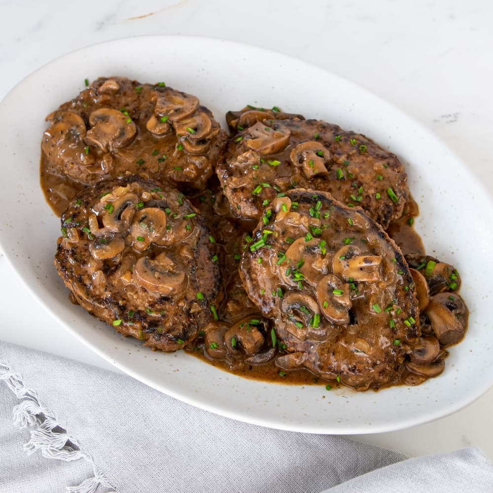 Four Salisbury steaks topped with mushroom gravy and chopped herbs are served on an oval white plate.