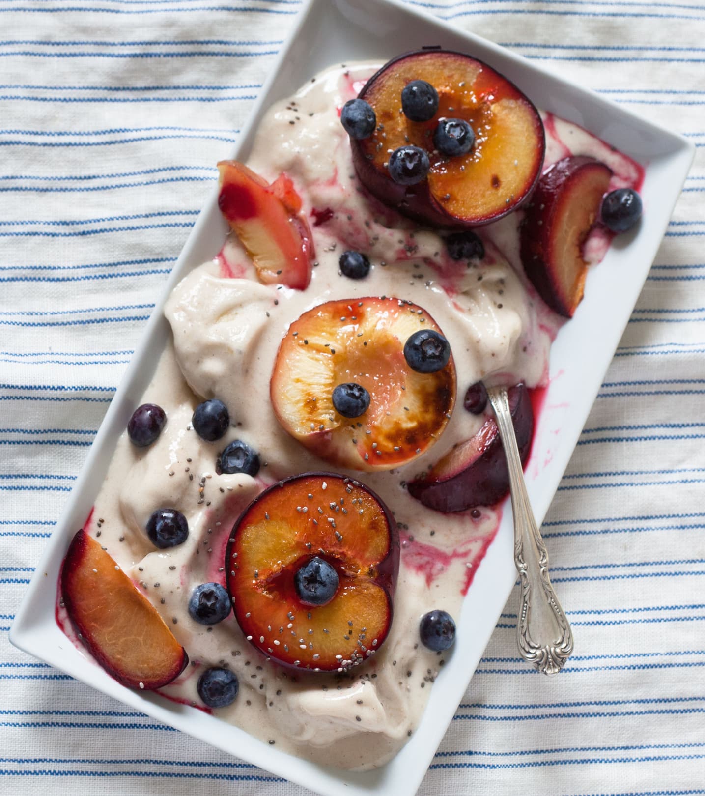 Rectangular dish of creamy dessert topped with halved plums, blueberries, chia seeds, and a spoon, on a striped cloth background.