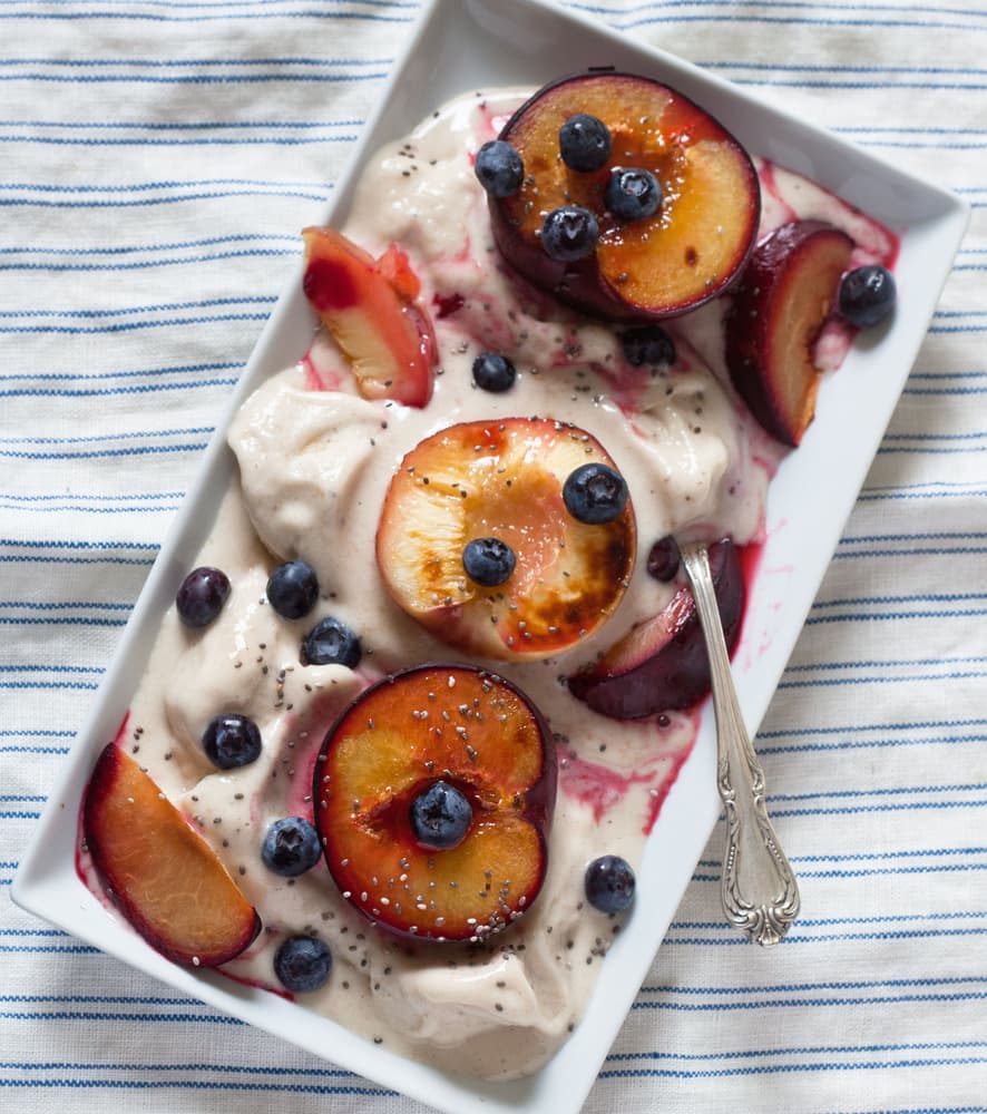 Rectangular dish of creamy dessert topped with halved plums, blueberries, chia seeds, and a spoon, on a striped cloth background.