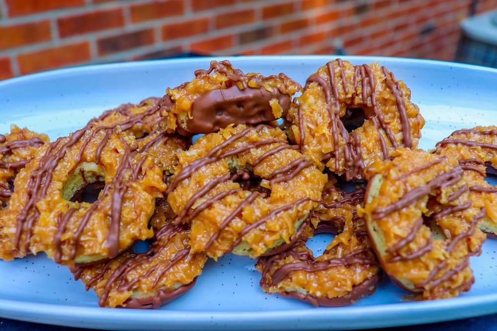 A plate of caramel and chocolate-drizzled cookies with a toasted coconut topping is displayed on a blue dish.