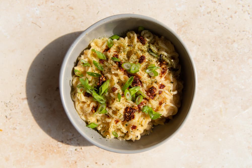 A bowl of cooked noodles topped with chopped green onions, chili oil, and seasoning, on a light-colored surface.