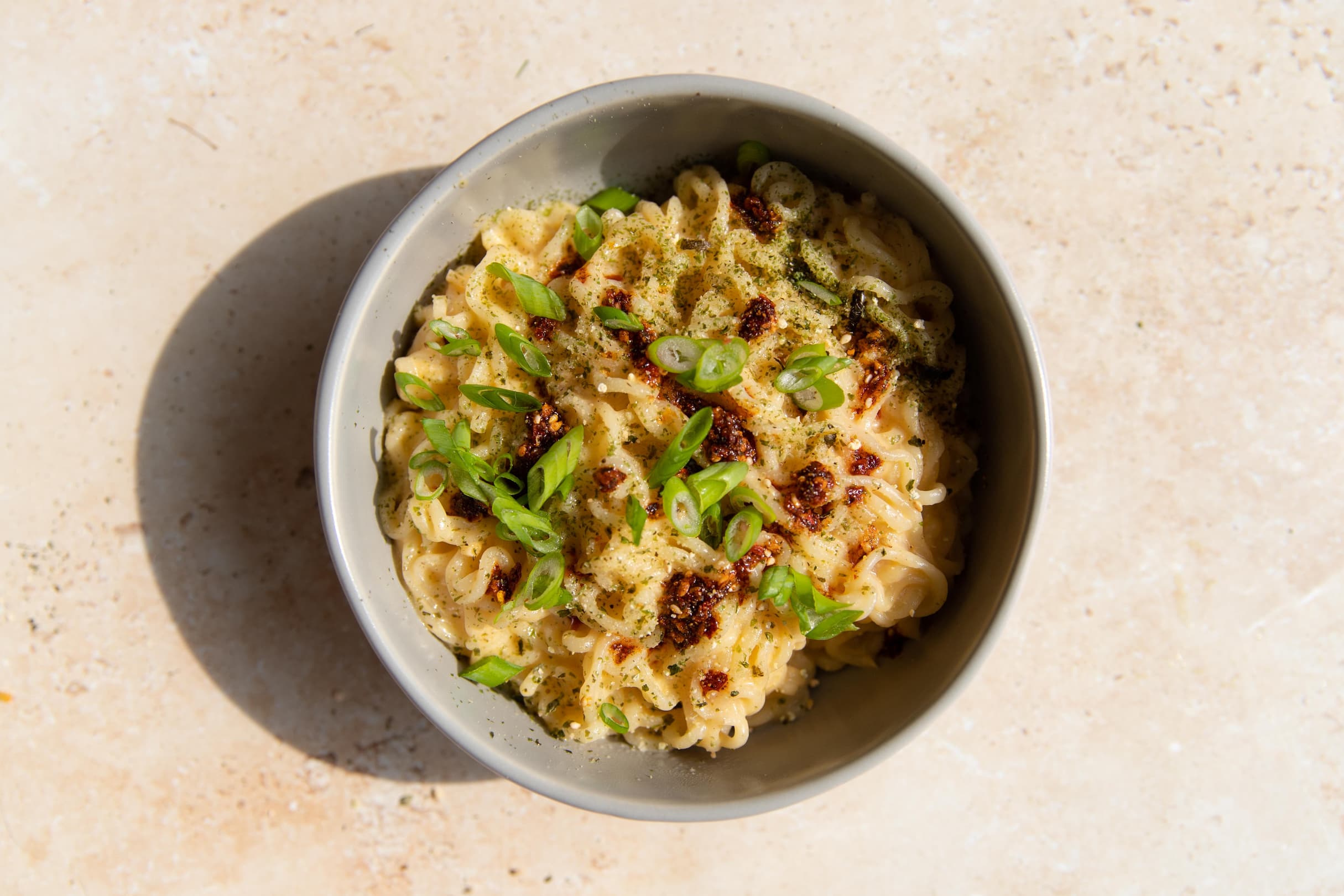 A bowl of cooked noodles topped with chopped green onions, chili oil, and seasoning, on a light-colored surface.