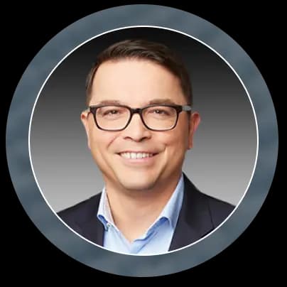 Smiling man wearing glasses, a light blue shirt, and a dark blazer, shown in a circular frame with a gray gradient background.