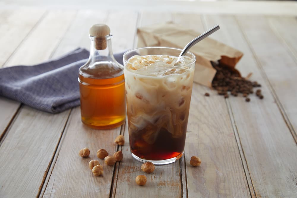 A glass of iced coffee with milk and a metal straw, surrounded by hazelnuts, a bottle of syrup, a cloth napkin, and a bag of coffee beans on a wooden table.