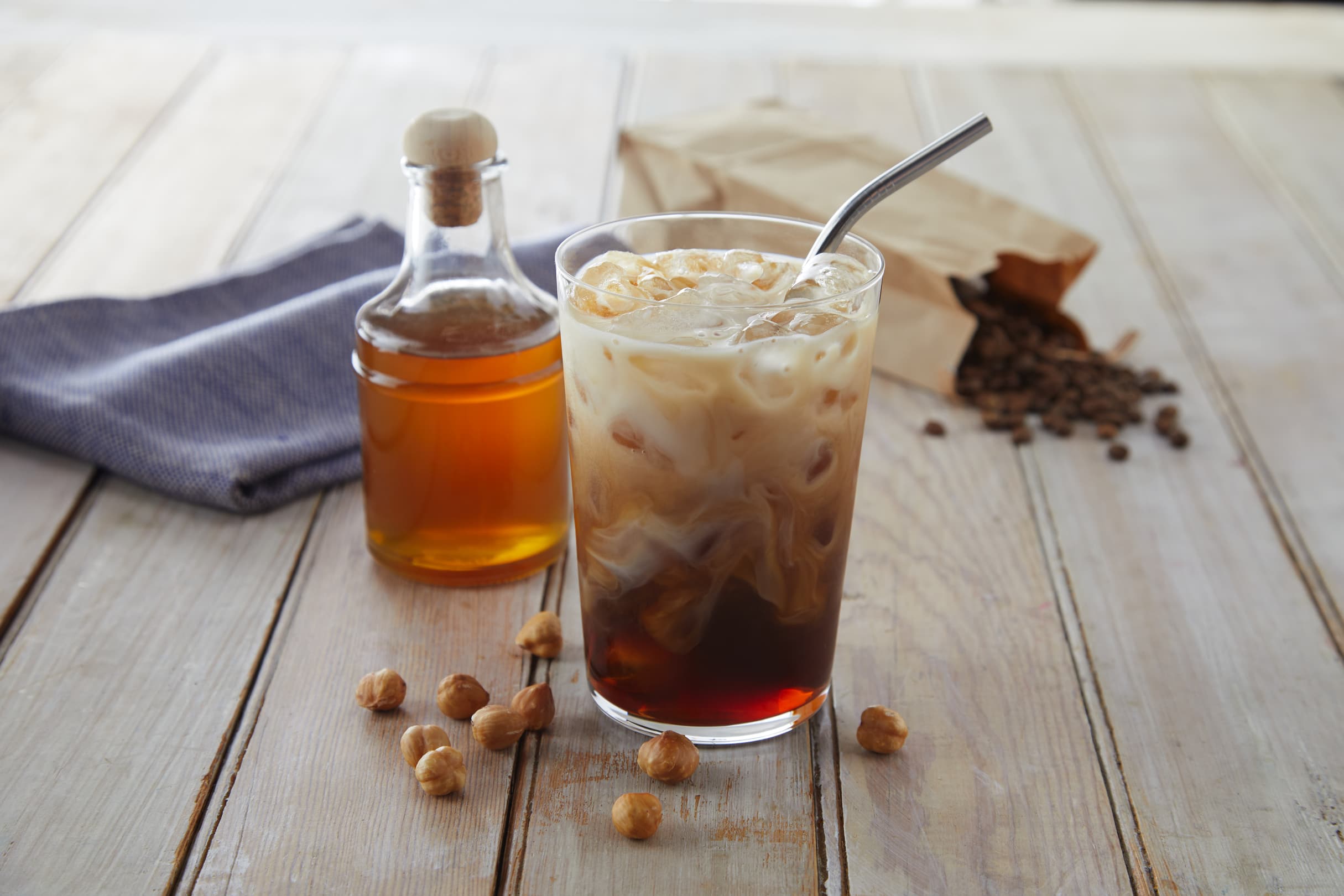 A glass of iced coffee with milk and a metal straw, surrounded by hazelnuts, a bottle of syrup, a cloth napkin, and a bag of coffee beans on a wooden table.
