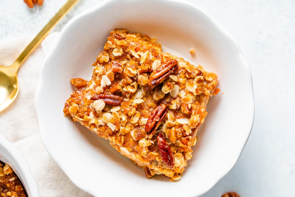 A square piece of pecan oat dessert sits in a white bowl, topped with oats and chopped pecans, beside a gold spoon.