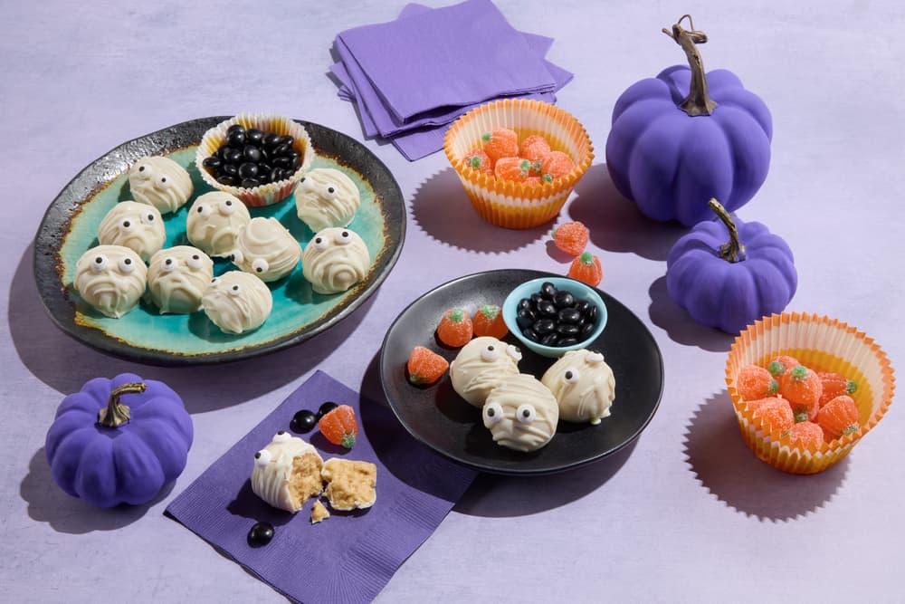 Plates of Halloween treats shaped like mummies, bowls of black and orange candy, and three purple decorative pumpkins on a lavender surface with purple napkins.