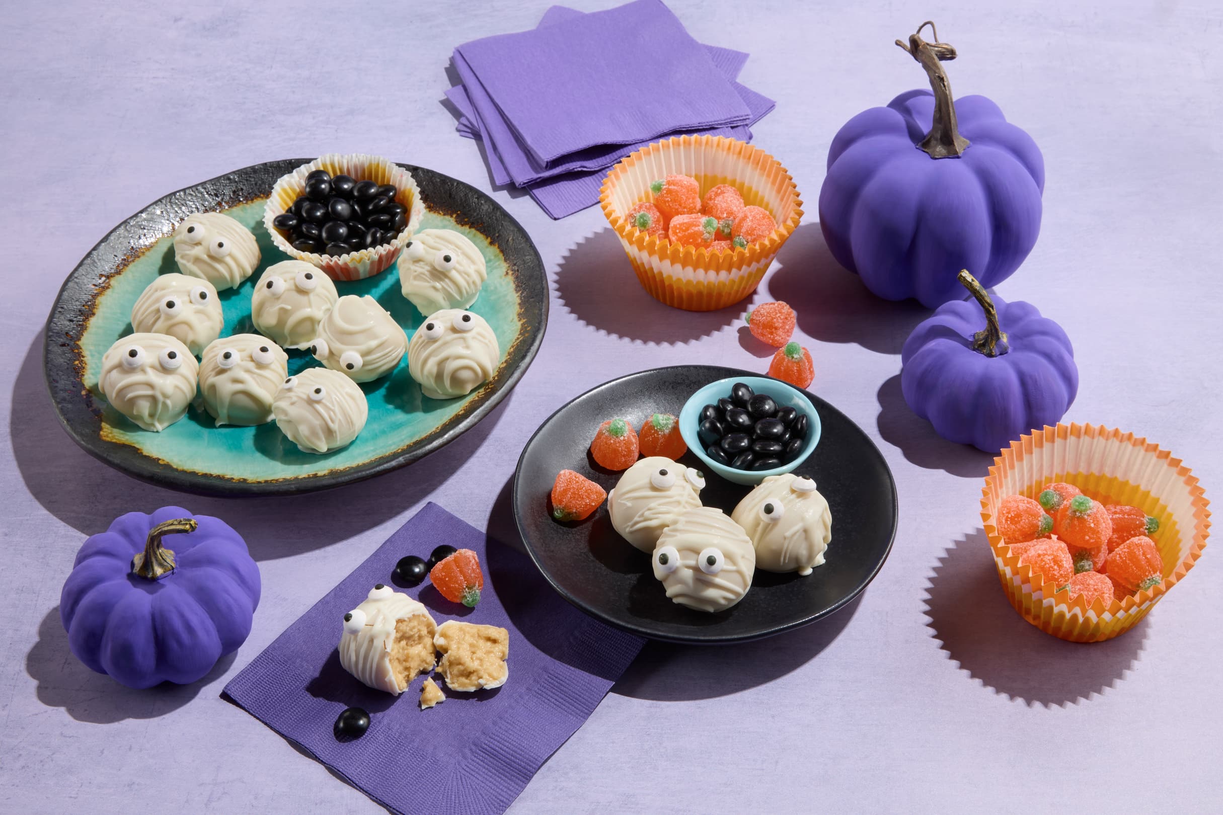 Plates of Halloween treats shaped like mummies, bowls of black and orange candy, and three purple decorative pumpkins on a lavender surface with purple napkins.