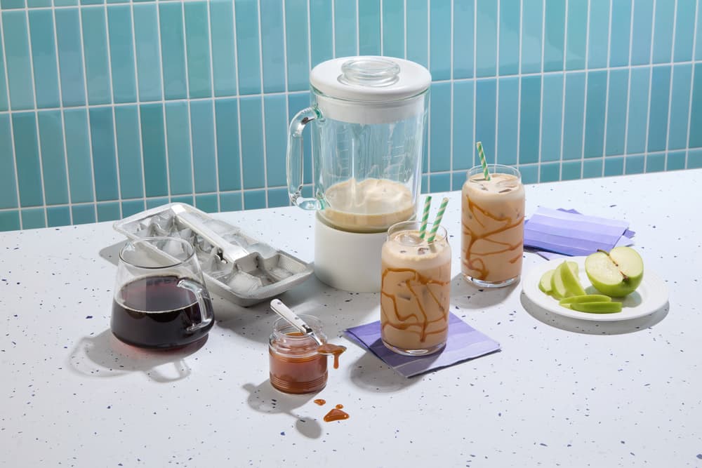 A blender, two iced coffee drinks with caramel drizzle, a glass pitcher, ice cube tray, caramel jar, jug of coffee, napkins, and sliced green apples on a speckled counter.