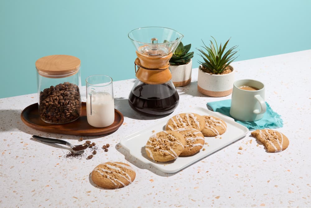 Cookies on a tray, a coffee carafe, mug, jar of coffee beans, milk, spoon, and small potted plants on a countertop with a blue background.