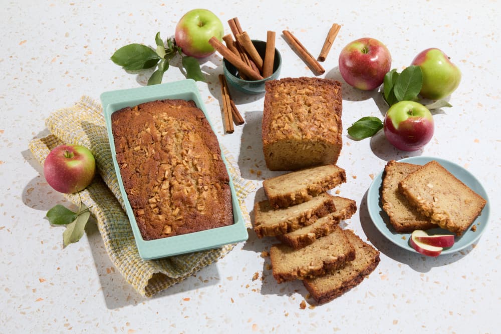 Sliced apple bread on a plate, a loaf in a baking pan, cinnamon sticks in a bowl, apples, and leaves arranged on a white surface.