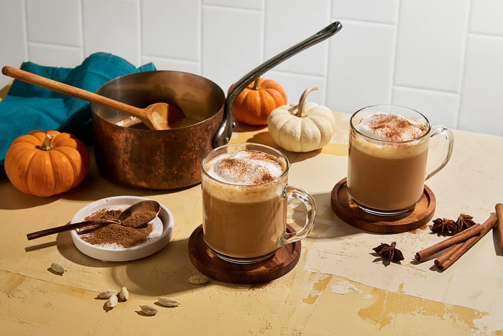 Two mugs of spiced latte on wooden coasters beside a pot, spices, spoons, and small pumpkins on a beige table with a white tiled background.