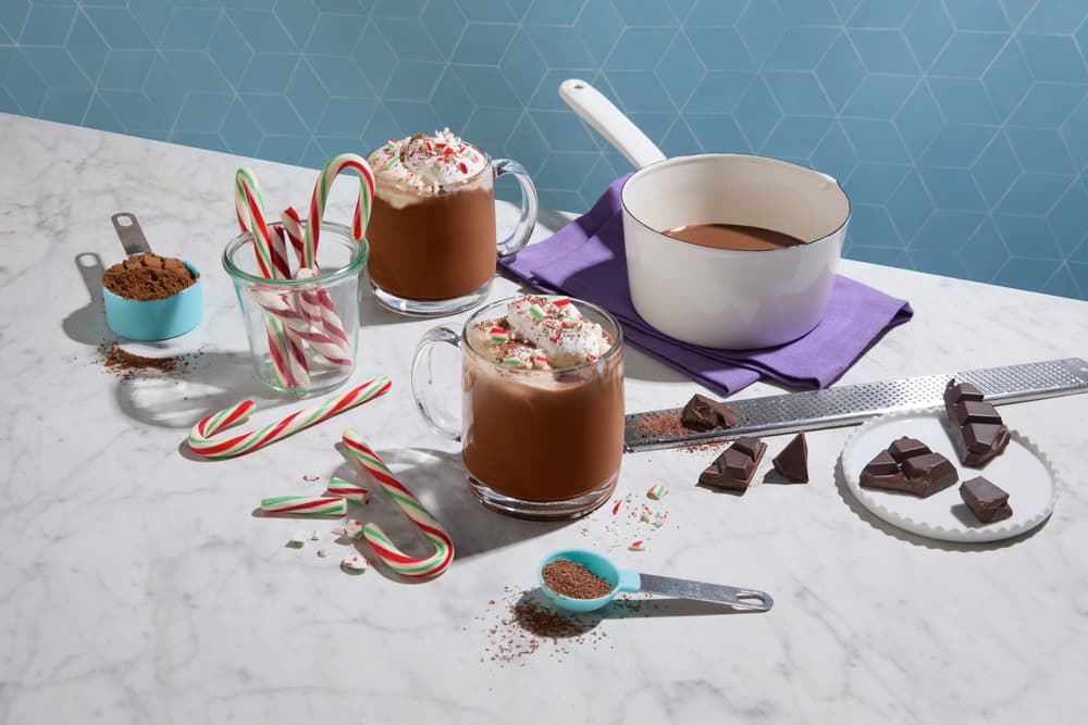 Two mugs of hot chocolate with whipped cream and crushed candy canes, surrounded by candy canes, cocoa powder, chocolate pieces, and a saucepan on a marble counter.