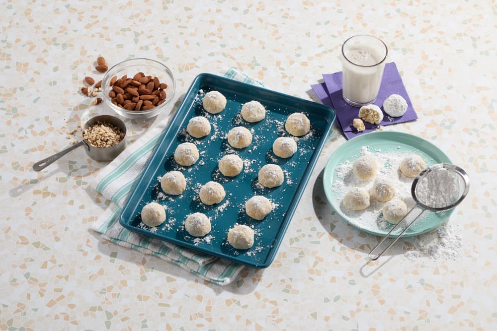 A blue tray with powdered sugar-coated cookies, a plate with more cookies being dusted, a glass of milk, almonds, chopped nuts, and a sifter on a patterned surface.