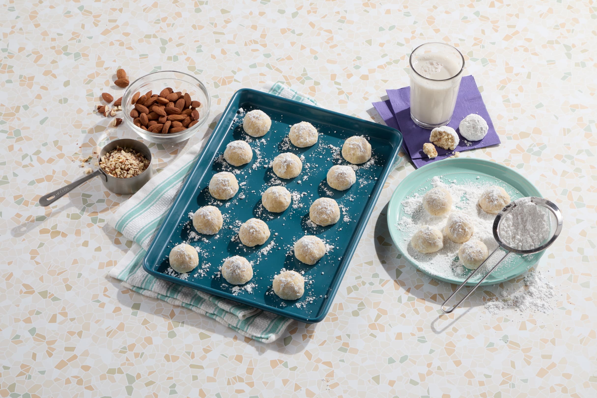 A blue tray with powdered sugar-coated cookies, a plate with more cookies being dusted, a glass of milk, almonds, chopped nuts, and a sifter on a patterned surface.