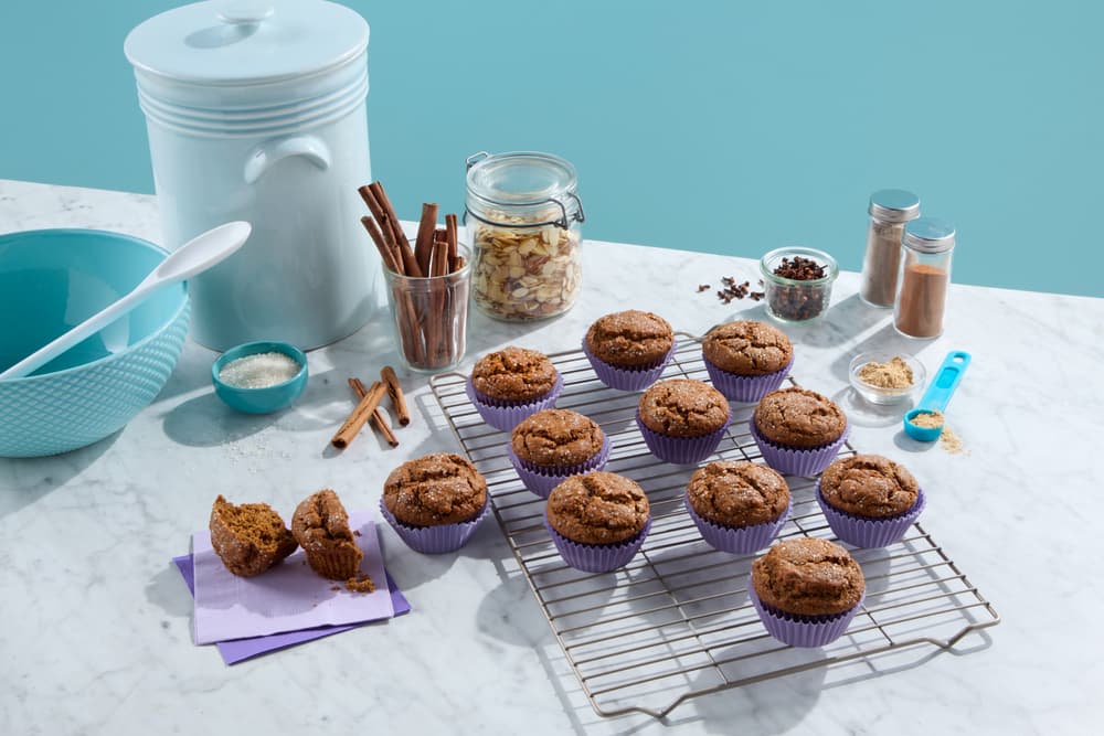 Twelve muffins on a cooling rack with baking ingredients, utensils, and jars of spices on a marble countertop against a blue background.