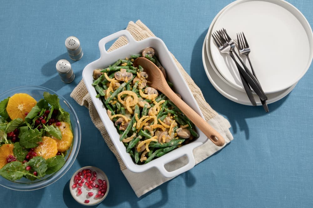 A casserole dish with green bean casserole, a wooden spoon, a salad with oranges and pomegranate, and a stack of plates with forks on a blue tablecloth.