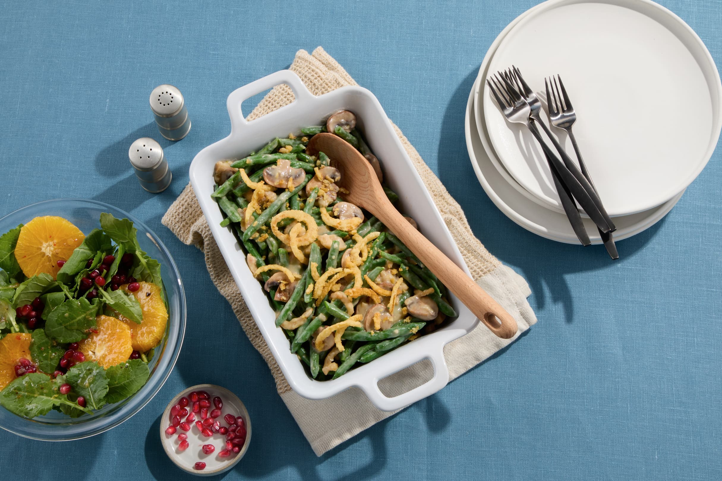 A casserole dish with green bean casserole, a wooden spoon, a salad with oranges and pomegranate, and a stack of plates with forks on a blue tablecloth.
