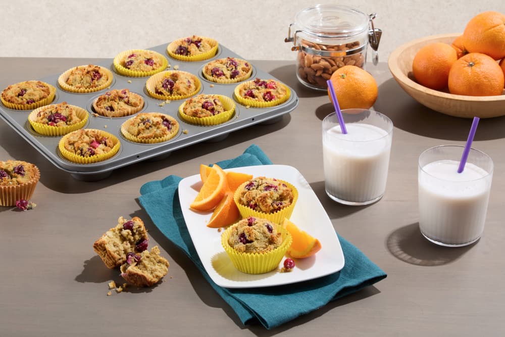A tray of muffins, a plate with a muffin and orange slices, two glasses of milk with straws, a bowl of oranges, and a jar of almonds on a table.