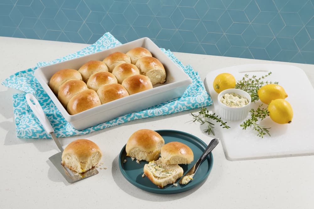 A baking dish with golden dinner rolls, some served on a blue plate. Nearby are lemons, thyme, a small bowl of butter, and a spatula, all on a white countertop.