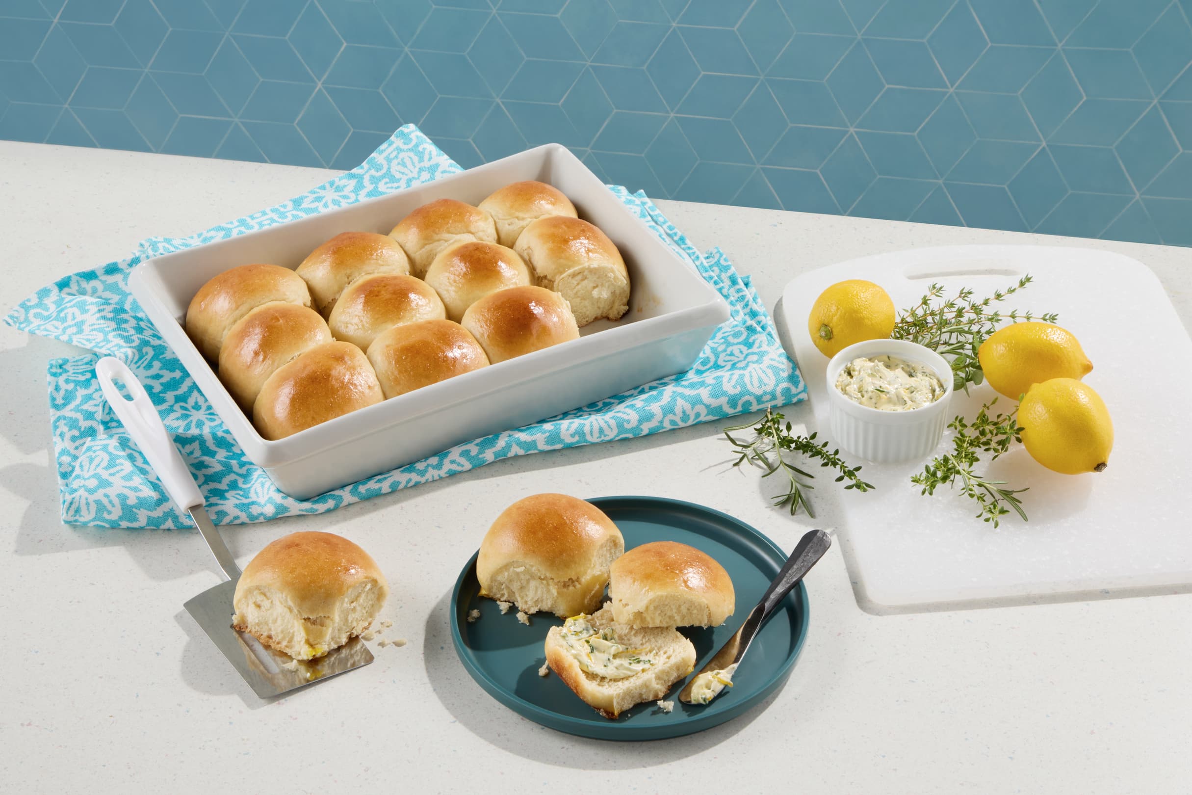 A baking dish with golden dinner rolls, some served on a blue plate. Nearby are lemons, thyme, a small bowl of butter, and a spatula, all on a white countertop.