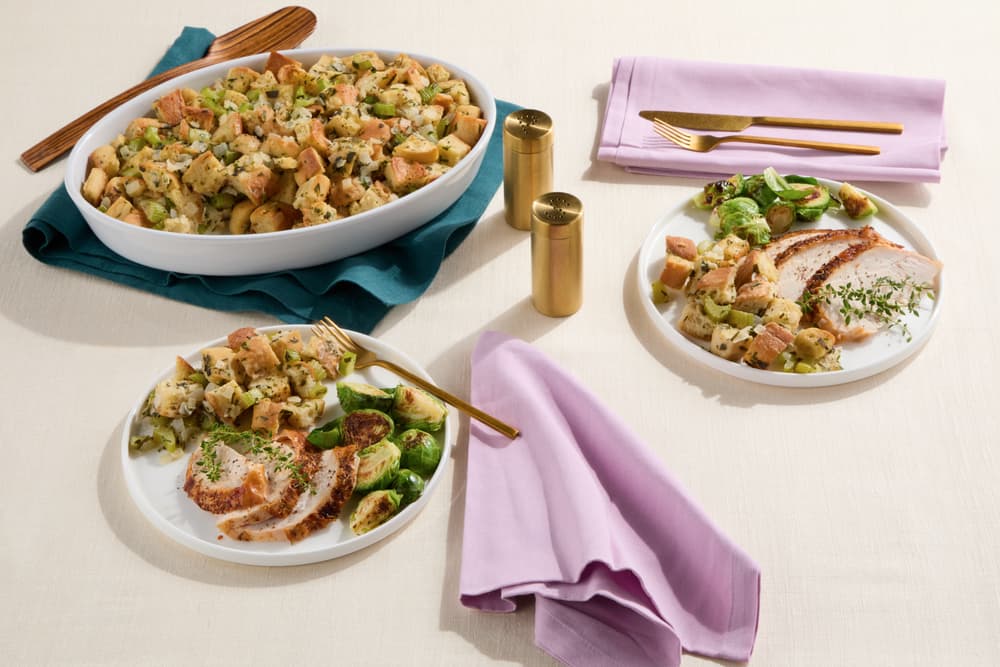 Two plates with sliced turkey, stuffing, and Brussels sprouts sit next to a large dish of stuffing, brass cutlery, and gold salt and pepper shakers on a cream tablecloth with pastel napkins.