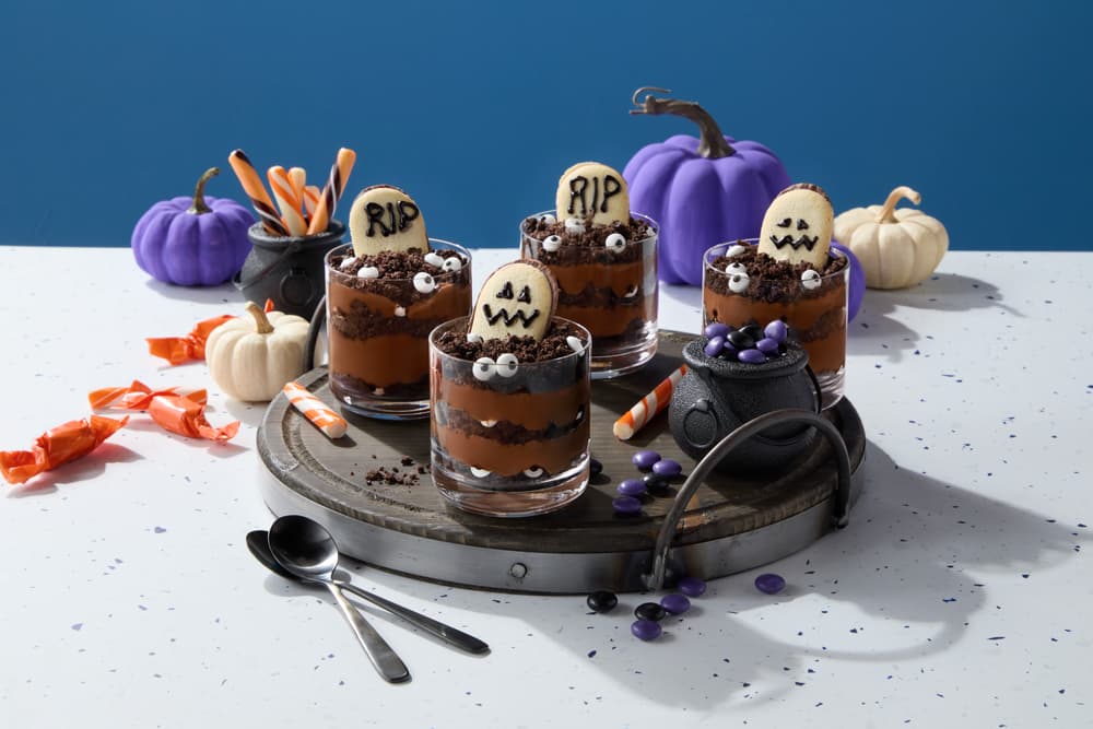 Five Halloween-themed chocolate pudding cups topped with cookie tombstones, candy eyeballs, and cauldrons filled with candy, are arranged on a round tray with pumpkins and spoons.