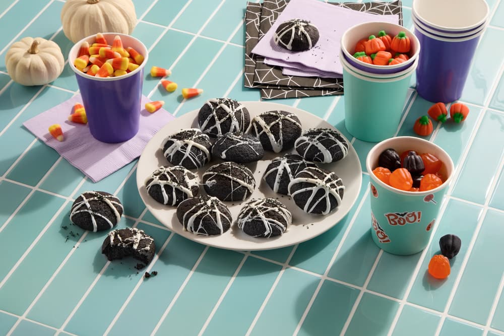 A plate of black cookies with white spiderweb icing sits on a tiled table with candy corn, gummy pumpkins, paper cups, napkins, and small decorative pumpkins.