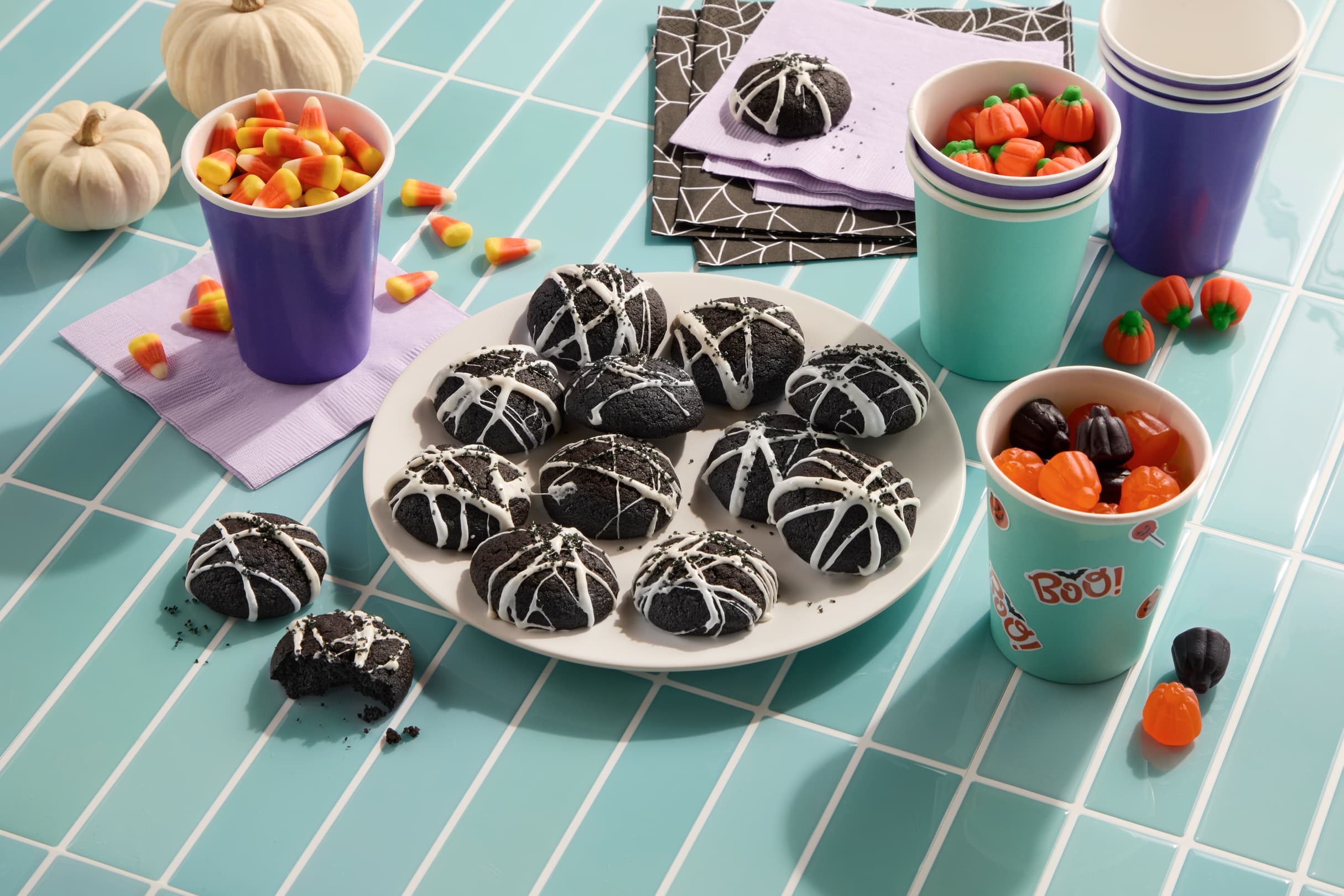 A plate of black cookies with white spiderweb icing sits on a tiled table with candy corn, gummy pumpkins, paper cups, napkins, and small decorative pumpkins.