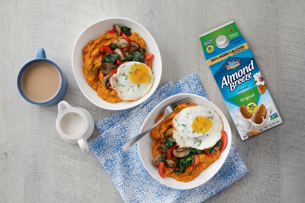 Two bowls of mashed sweet potatoes topped with fried eggs, sautéed vegetables, and herbs, next to a cup of coffee, a milk jug, and a carton of Almond Breeze almond milk on a grey surface.