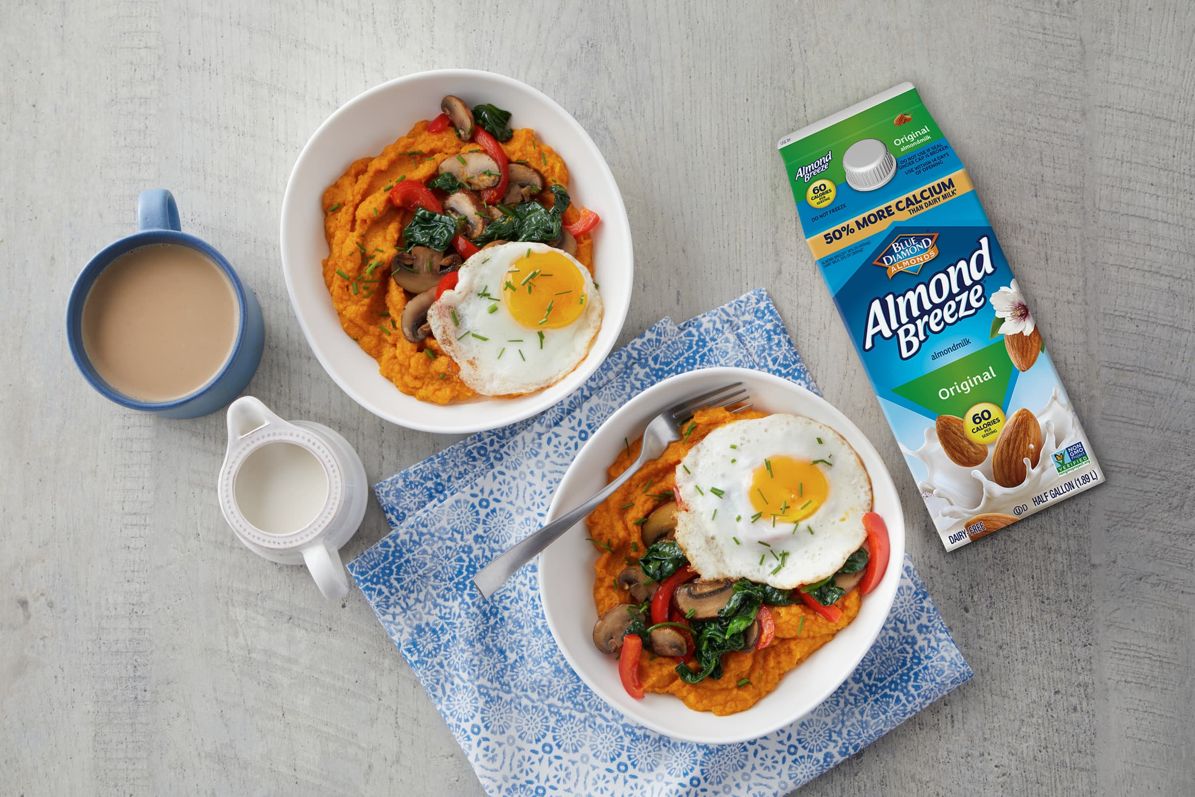 Two bowls of mashed sweet potatoes topped with fried eggs, sautéed vegetables, and herbs, next to a cup of coffee, a milk jug, and a carton of Almond Breeze almond milk on a grey surface.
