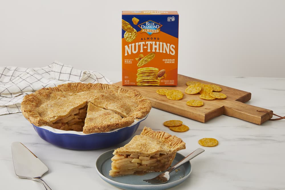 A sliced apple pie sits in a blue dish with a pie server nearby, a slice on a plate with a fork, and a box of Nut-Thins crackers and crackers spread on a wooden board in the background.