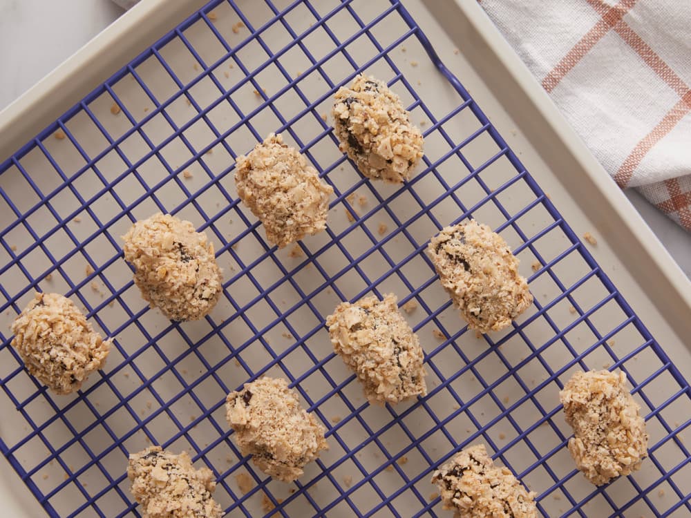 Oval-shaped cookies coated with chopped nuts are arranged on a blue cooling rack placed over a baking sheet, with a checkered cloth partially visible in the corner.