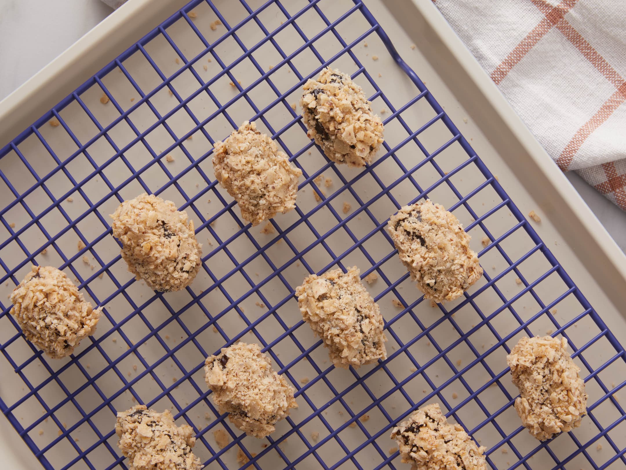 Oval-shaped cookies coated with chopped nuts are arranged on a blue cooling rack placed over a baking sheet, with a checkered cloth partially visible in the corner.