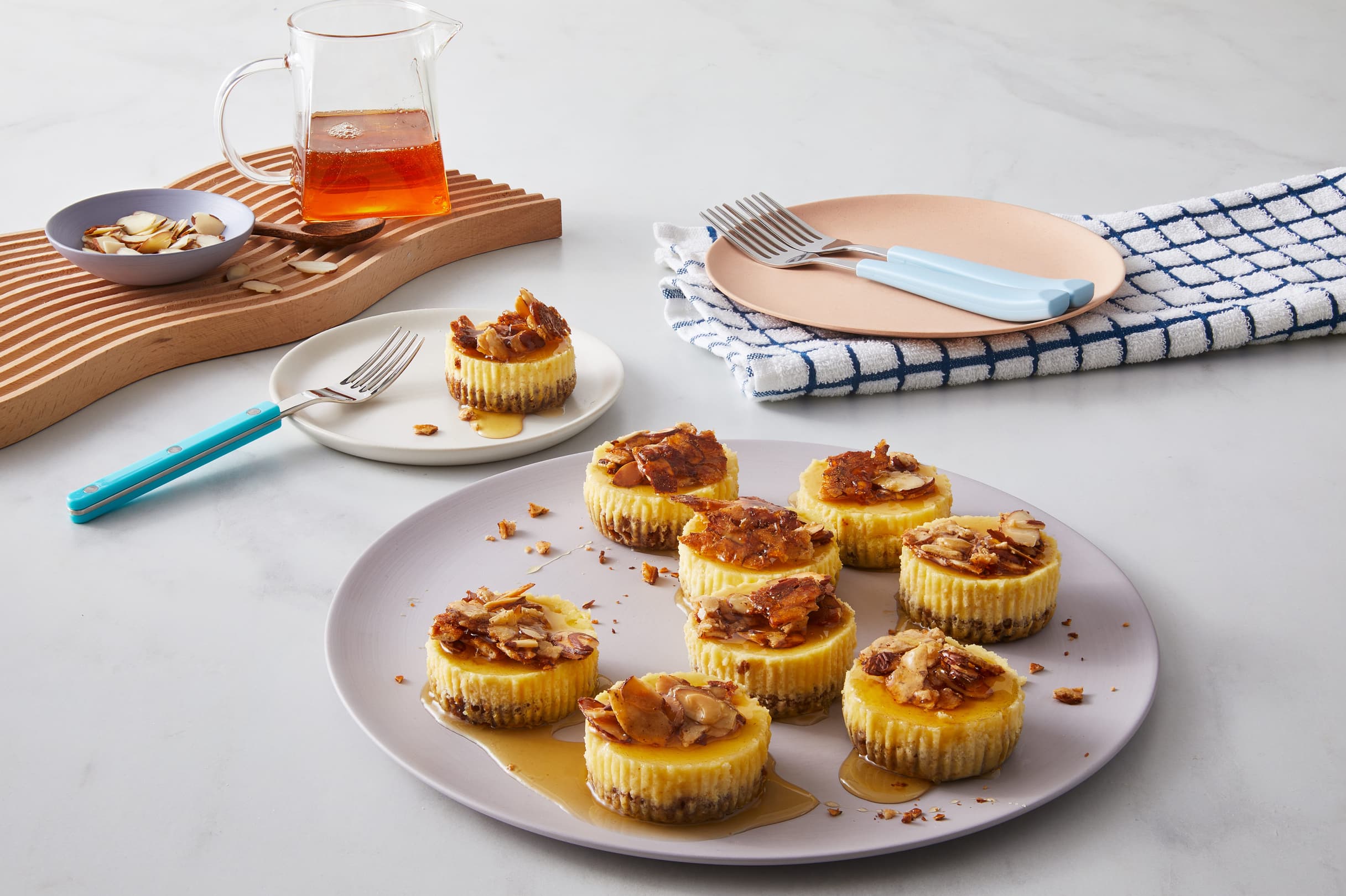 A plate of mini cheesecakes topped with nuts and syrup, surrounded by plates, forks, a bowl of sliced almonds, and a pitcher of syrup on a white surface.