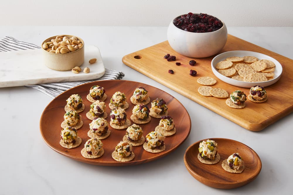 Round crackers topped with a creamy spread, herbs, and dried fruit on wooden plates, next to a bowl of nuts, dried cranberries, and plain crackers.