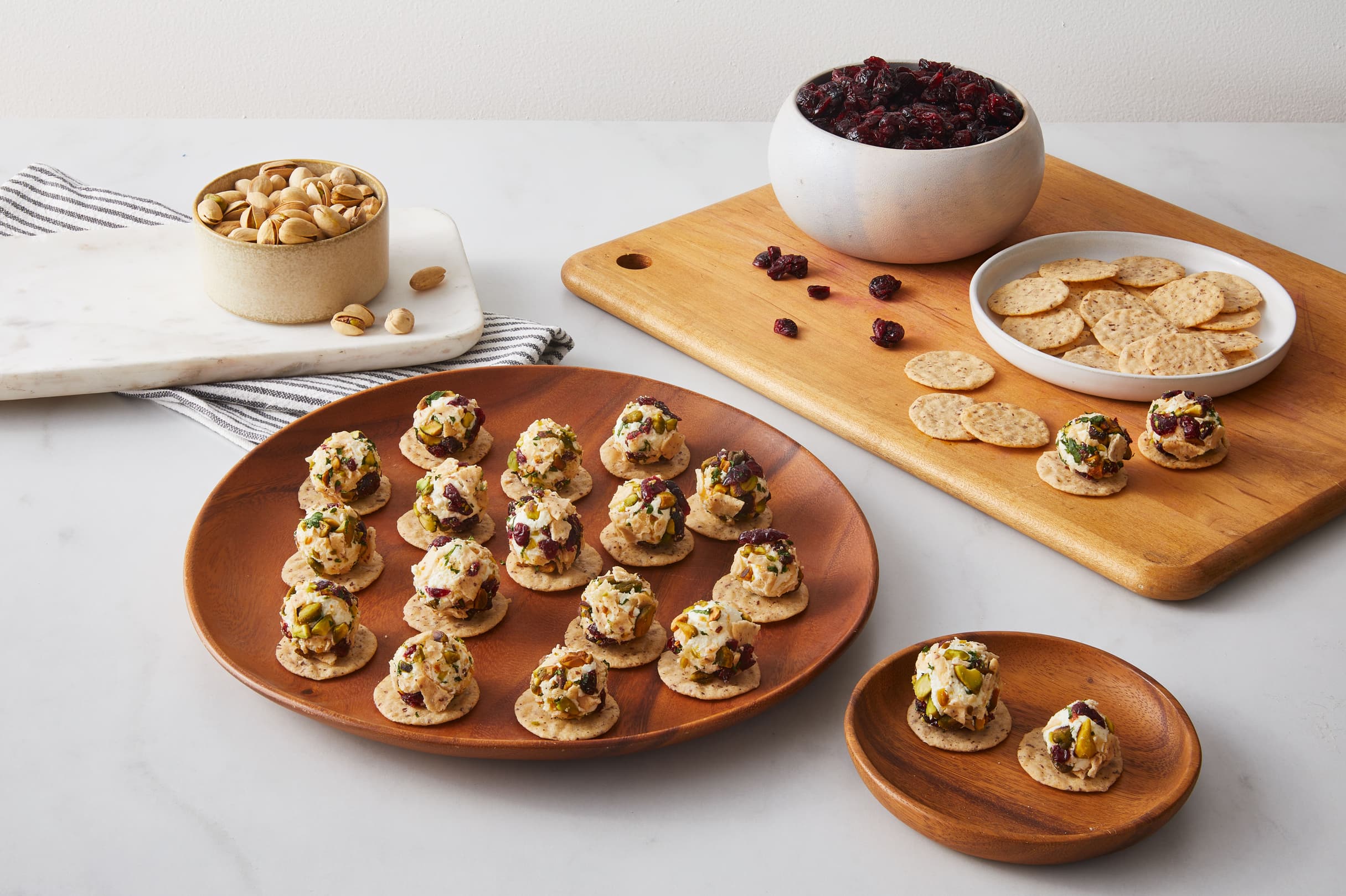Round crackers topped with a creamy spread, herbs, and dried fruit on wooden plates, next to a bowl of nuts, dried cranberries, and plain crackers.