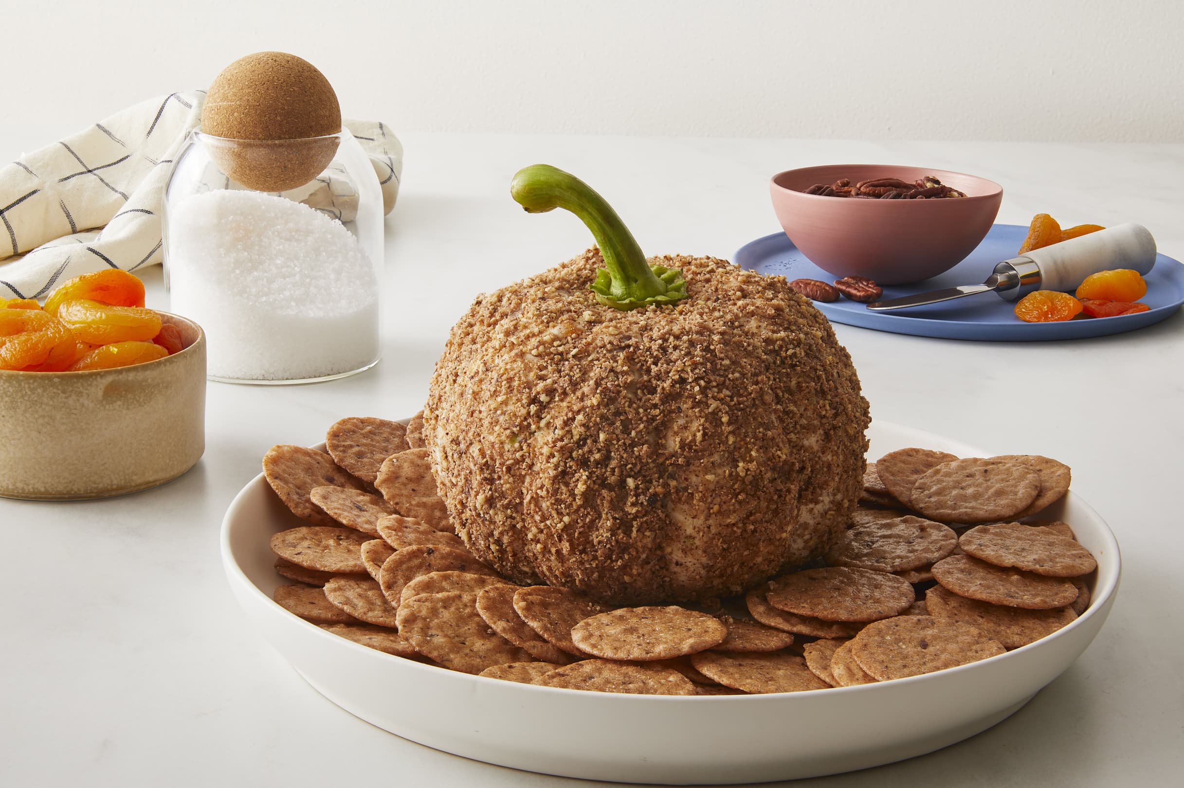 A cheese ball shaped like a pumpkin with a stem, coated in nuts, is served on a plate with round crackers. Bowls of dried fruit, pecans, and a jar of salt are in the background.