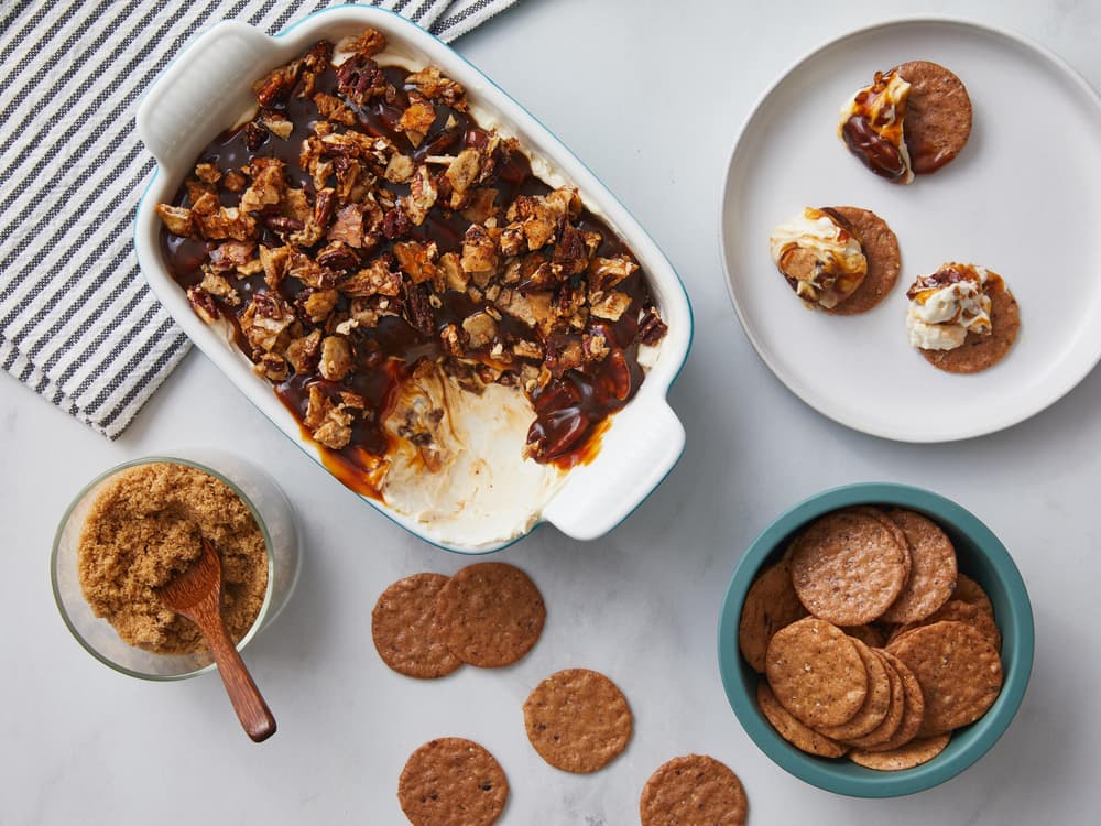 A casserole dish with a layered dip topped with chips, surrounded by round crackers, a bowl of brown sugar, and a striped napkin on a marble surface.
