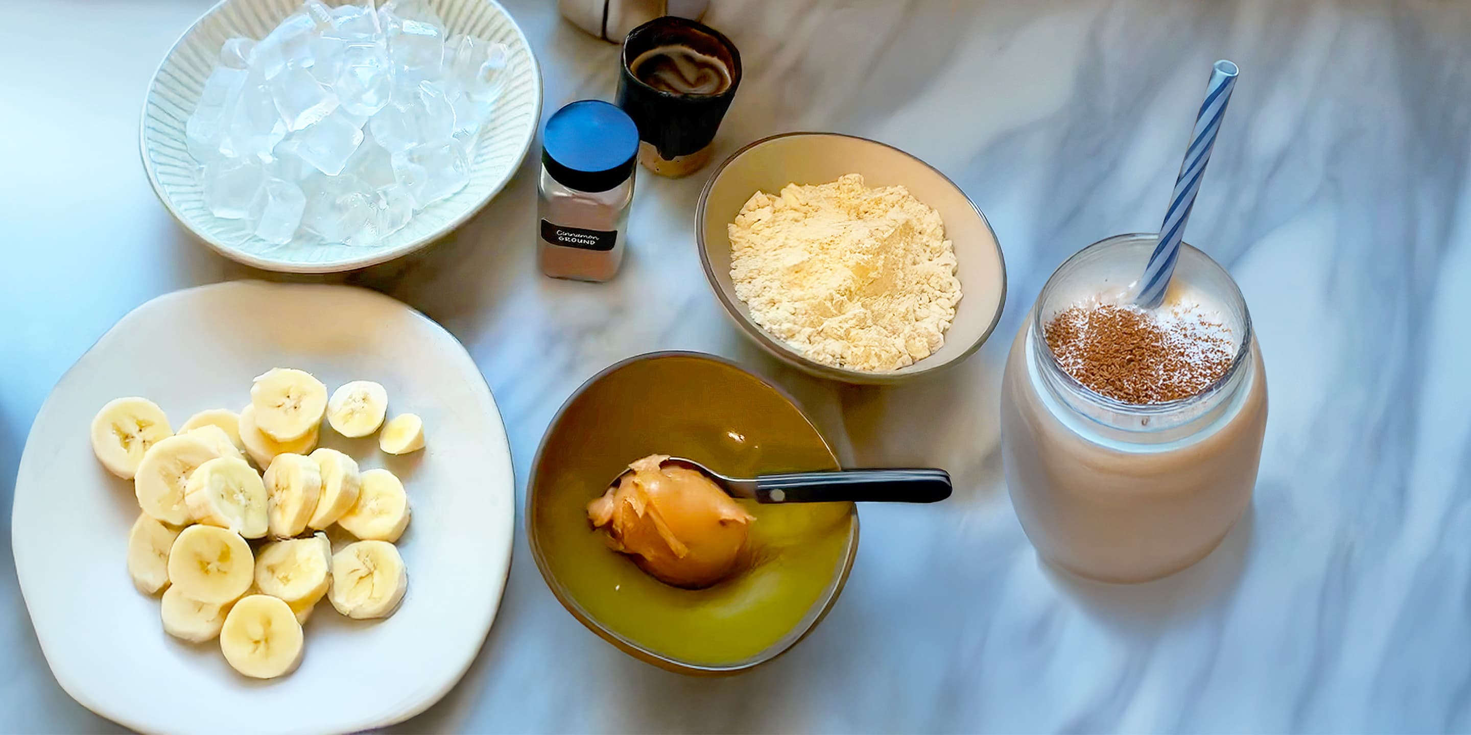 A bowl of ice, sliced banana, peanut butter, protein powder, cinnamon, cocoa powder, and a glass of smoothie with a straw are arranged on a marble surface.